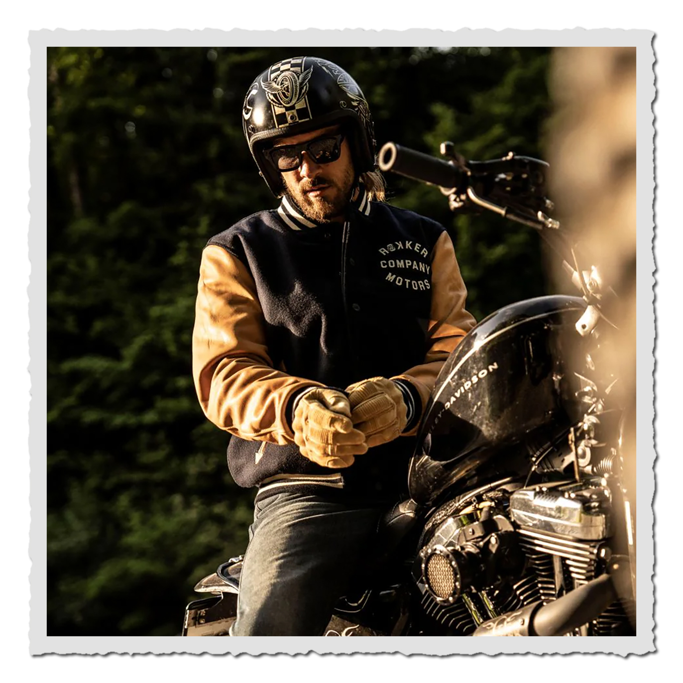 Motorradfahrer in Helm sitzt am Bike, trägt navyfarbene Jacke mit braunen Ärmeln, Abendlicht, unscharfer Wald im Hintergrund.